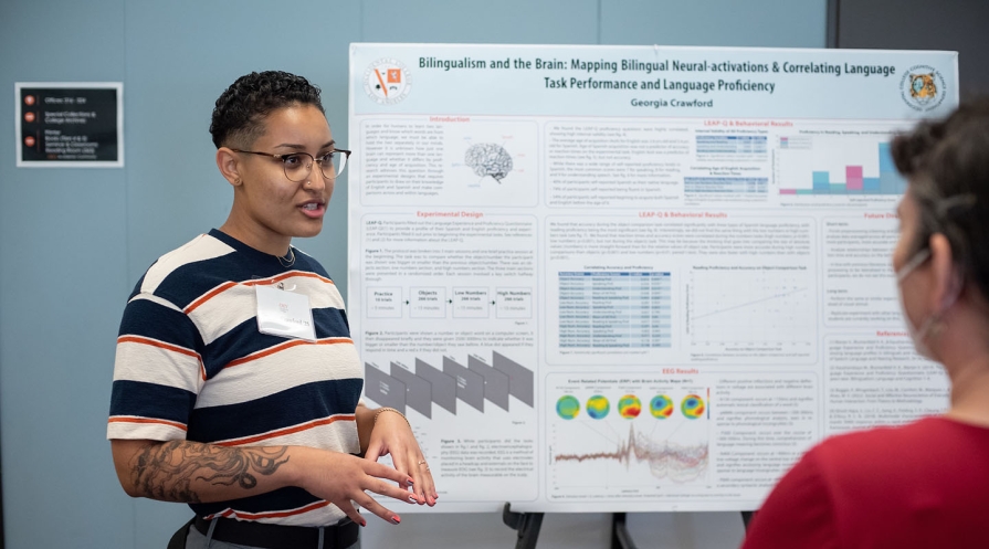 a student stands next to their research poster explaining the content to an onlooker