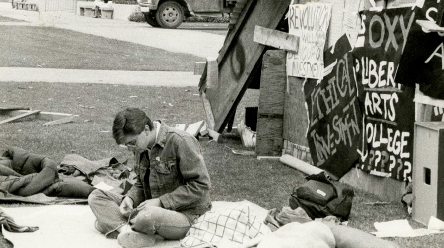 A black and white photo of an Oxy student sitting in protest with a number of signs in the background