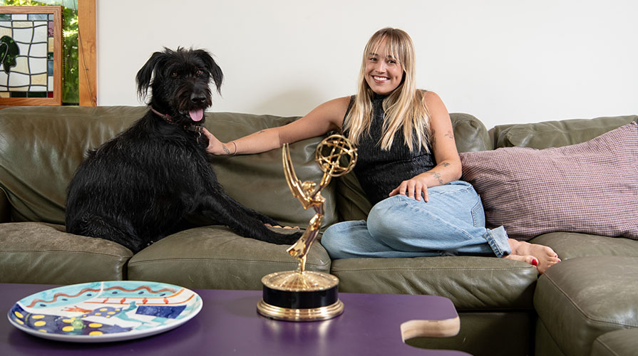 a young woman with long blonde hair sits with her black dog on a couch in front of her Emmy award