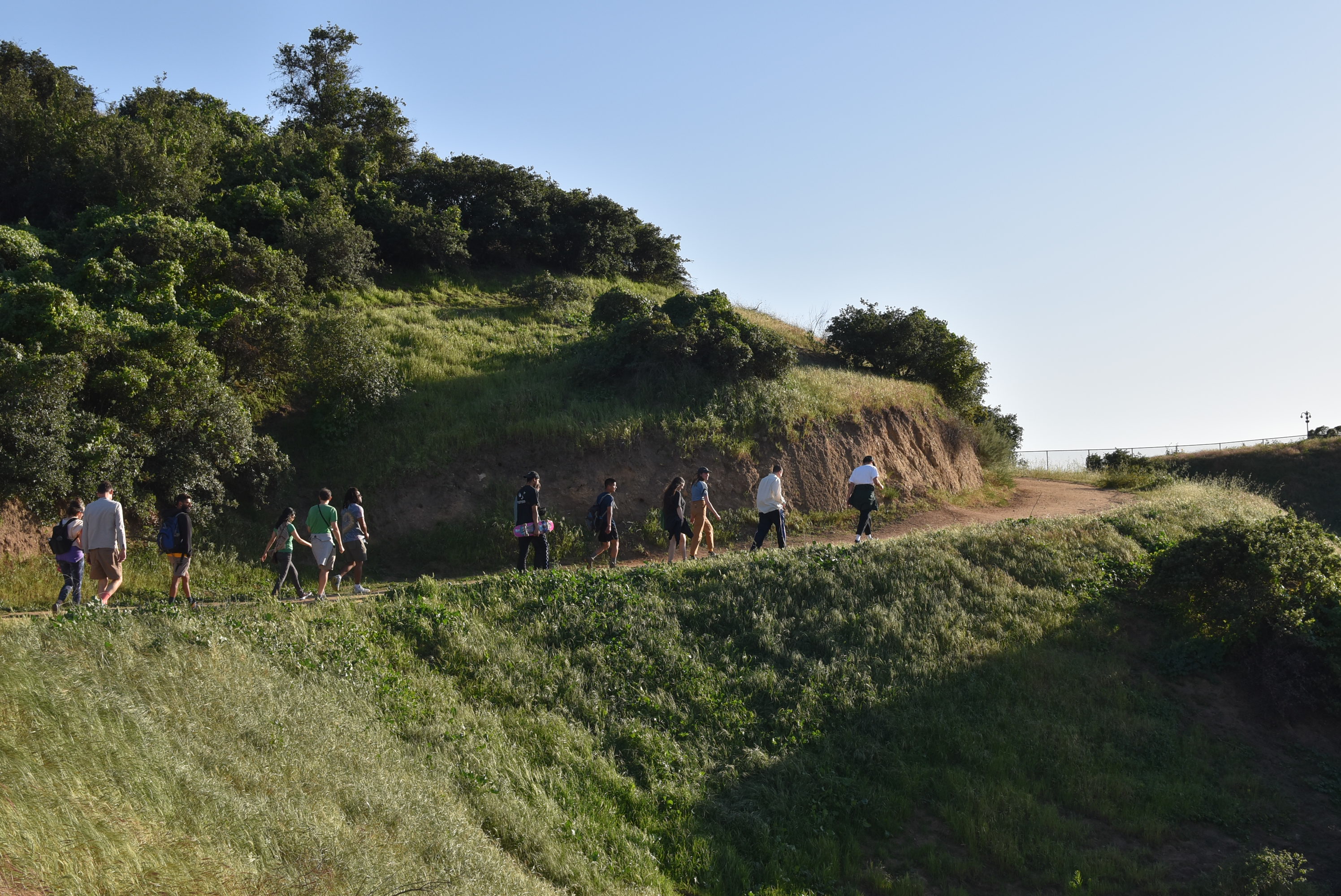 A wide image photograph of a group of people walking along a dirt path on a green hillside. 