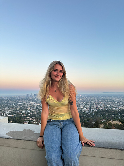 Grace is sitting on a wall with the city and sunsetting sky in background