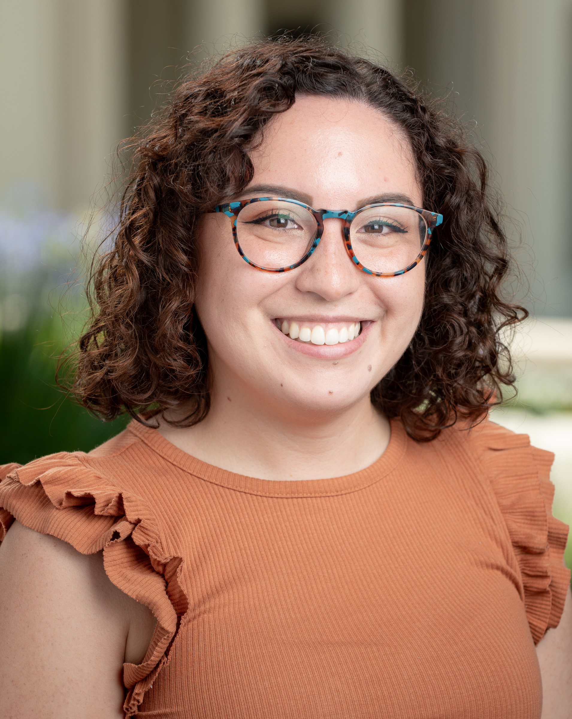 Image displays a woman with shoulder-length brunette curly hair and glasses wearing an orange shirt