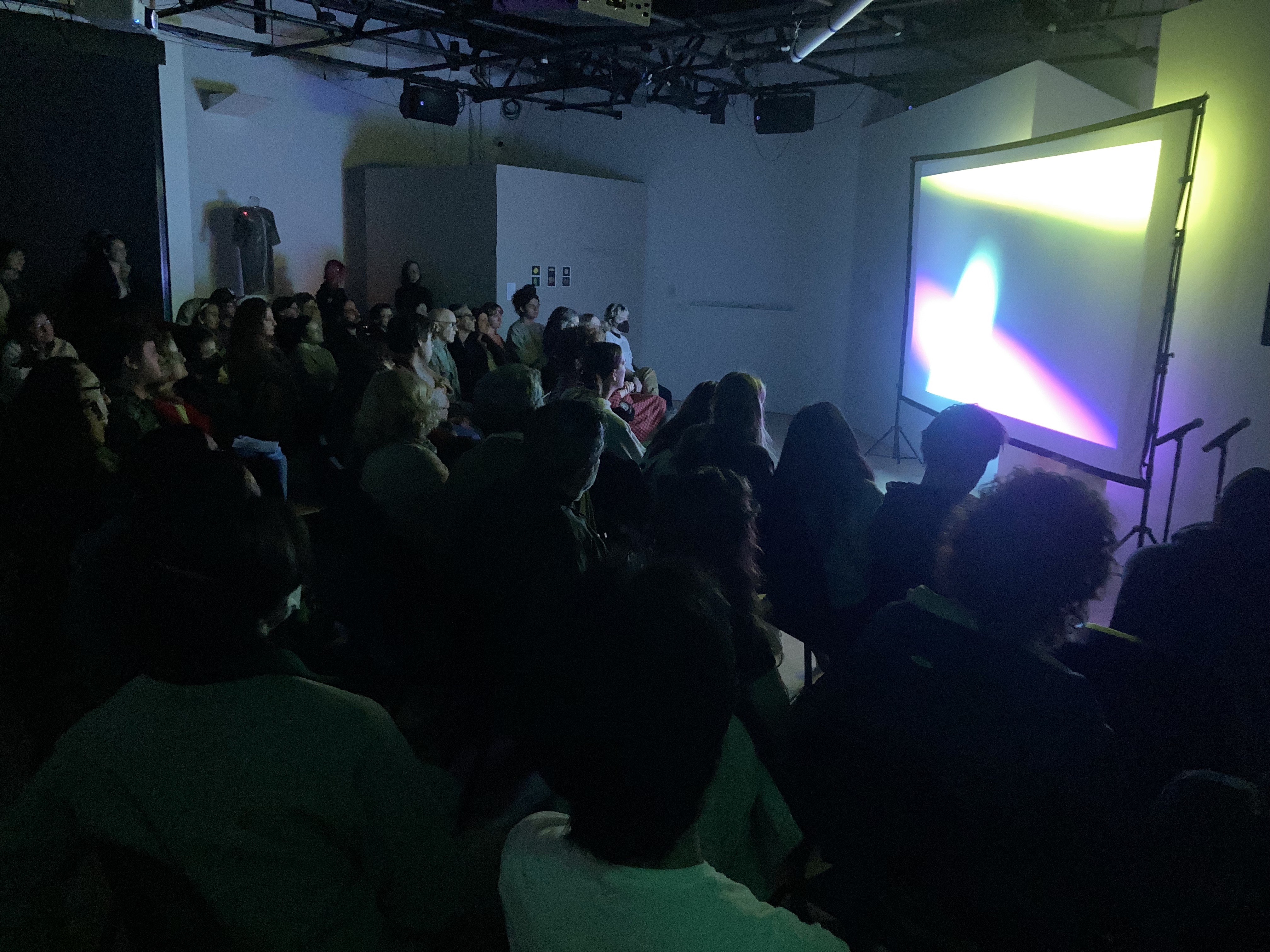 A group of people is seated watching a screening in the OXY ARTS gallery. The room is dark and the screen is lit with bright abstract colors. 