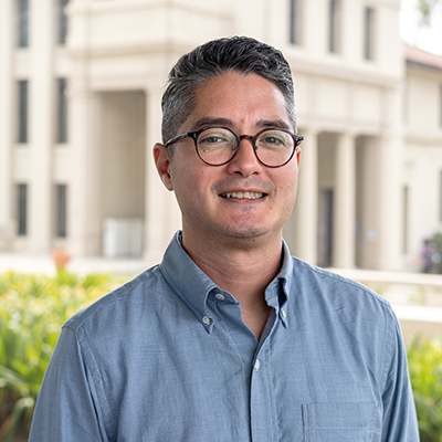 Joel is wearing a blue shirt and is standing in front of a building at the Occidental College campus