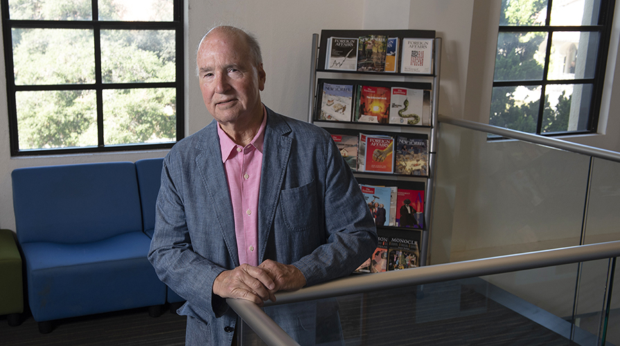Derek Shearer in a blue blazer in front of a bookshelf and a window on campus