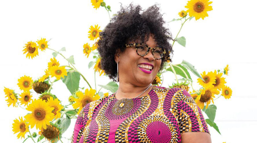 Monica White headshot in a colorful dress in front of tall sunflower plants