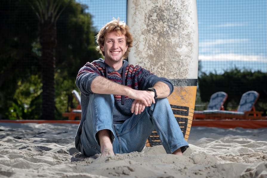 occidental student posing for photo while sitting in sand with a surfboard in the background