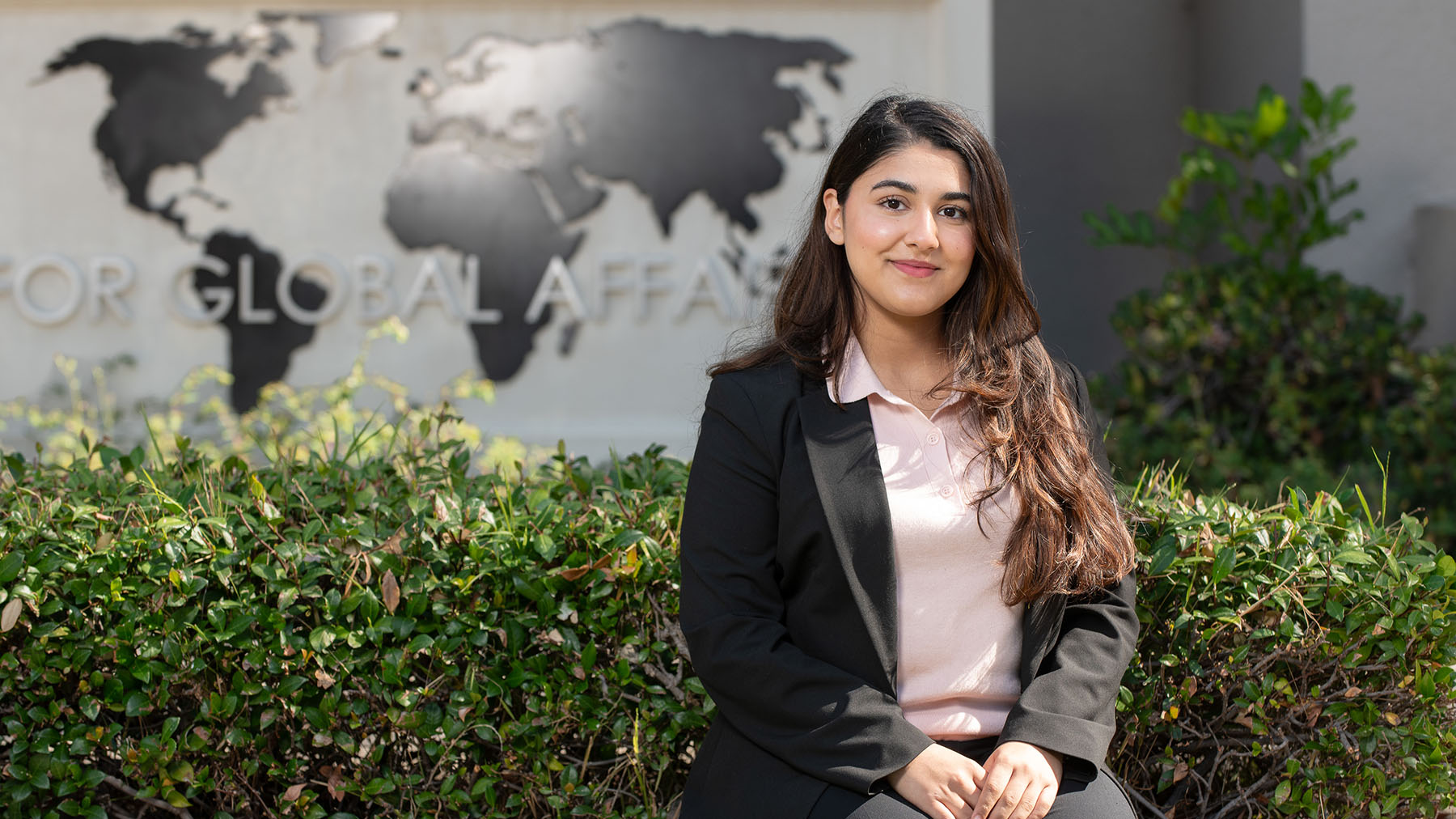 a young Occidental College student with long brown hair and a blazer seated in front of a bush outdoors