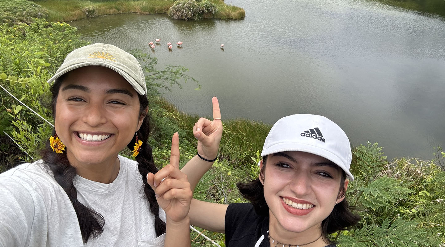 Two Occidental College students posing together in the Galapagos Islands pointing at some pink flamingos