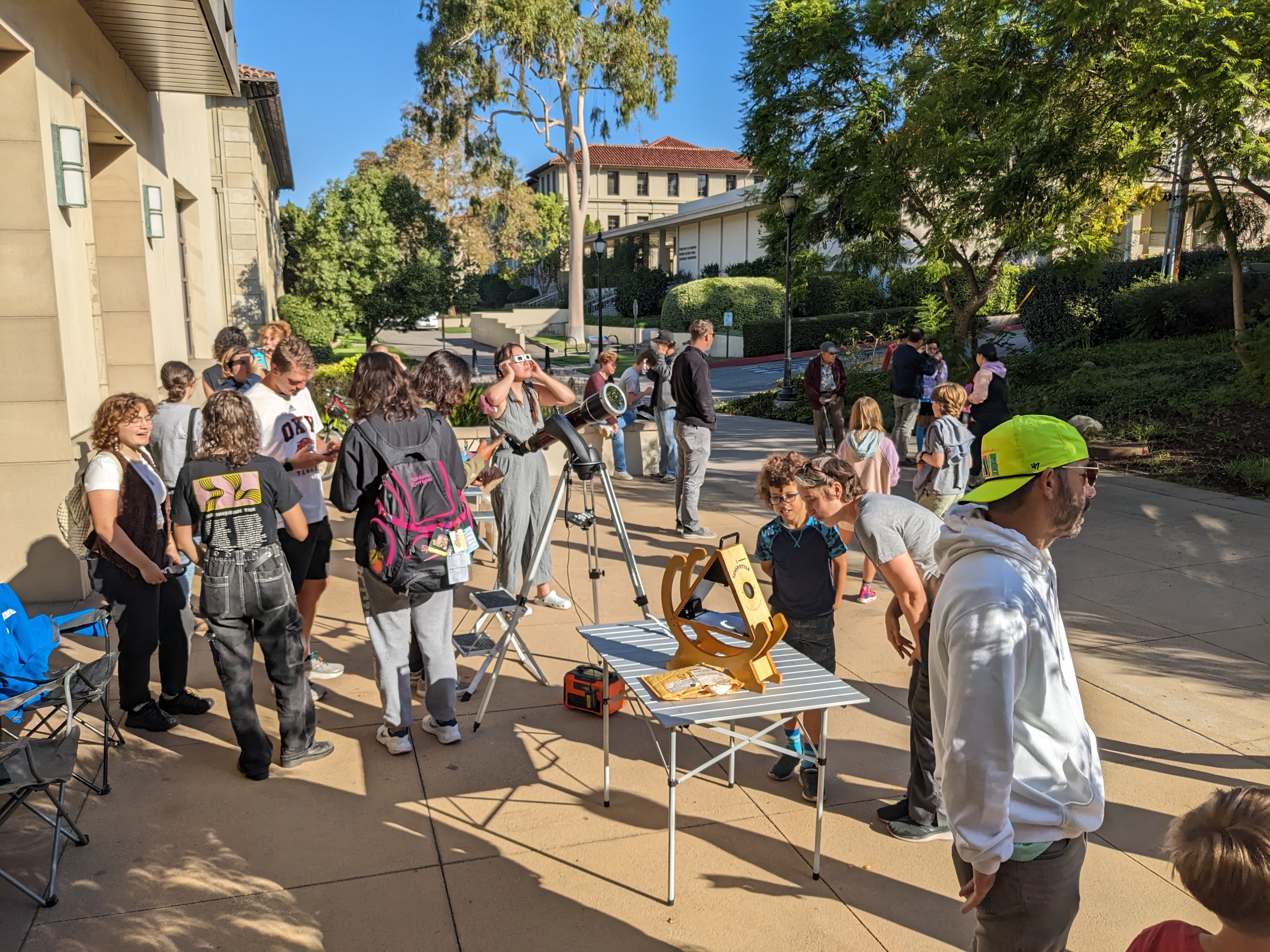 Oxy faculty, staff, students, and members of the local community standing around a telescope and sunspotter. Some are wearings solar eclipse glasses and looking at the partial solar eclipse.