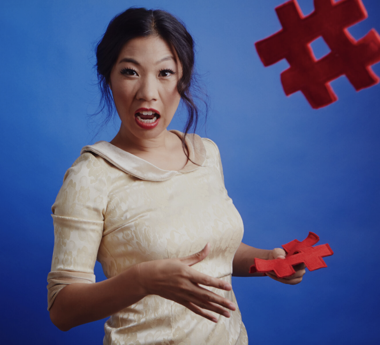 Screen Shot 2022-09-20 at 1.18.16 PM.png Portrait of Kristina Wong in a white dress standing in front of a bright blue background. She is throwing a red sculptural shape towards the camera.