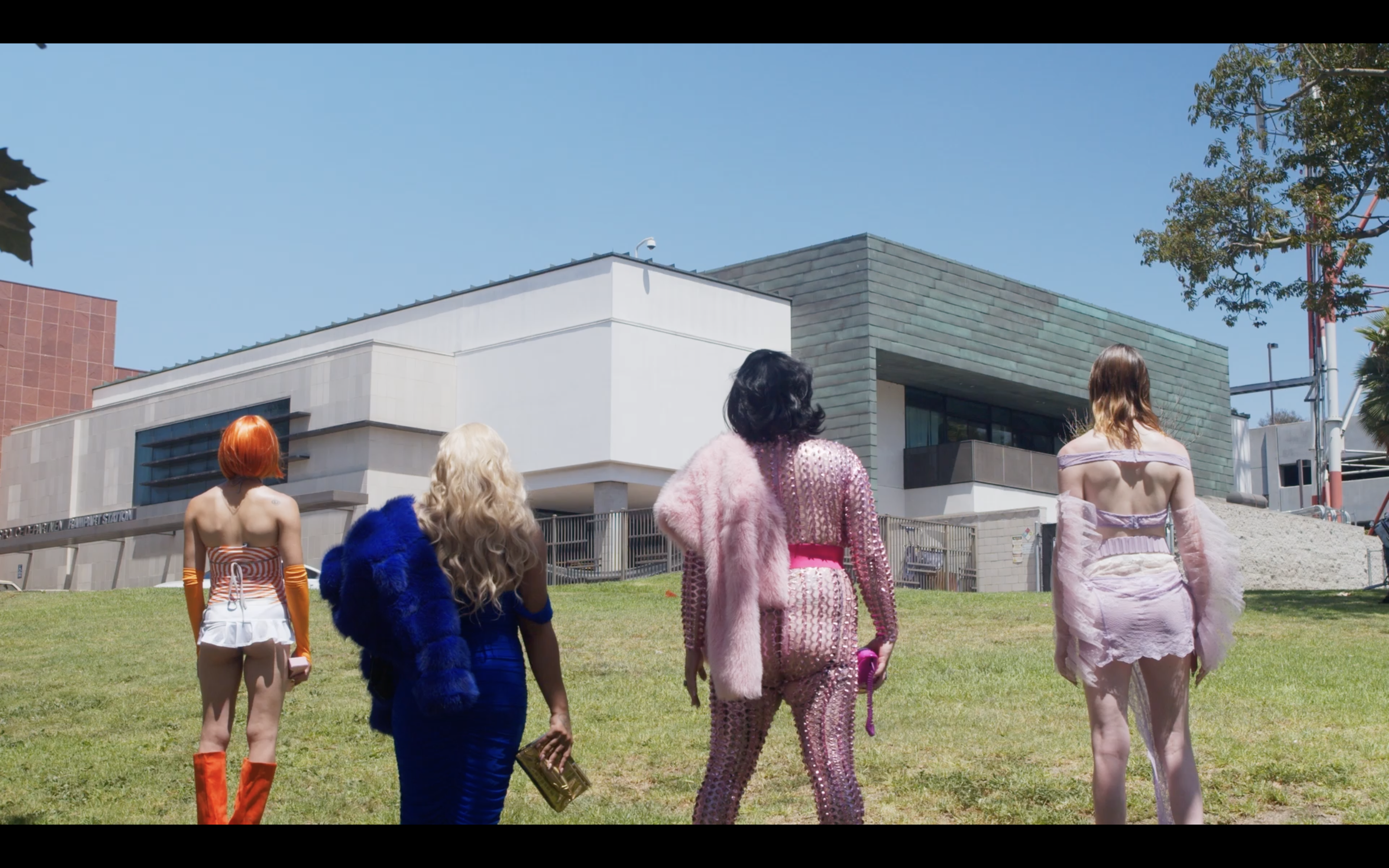 A still image of performers in vibrant, avant-garde outfits standing in front of a building. 