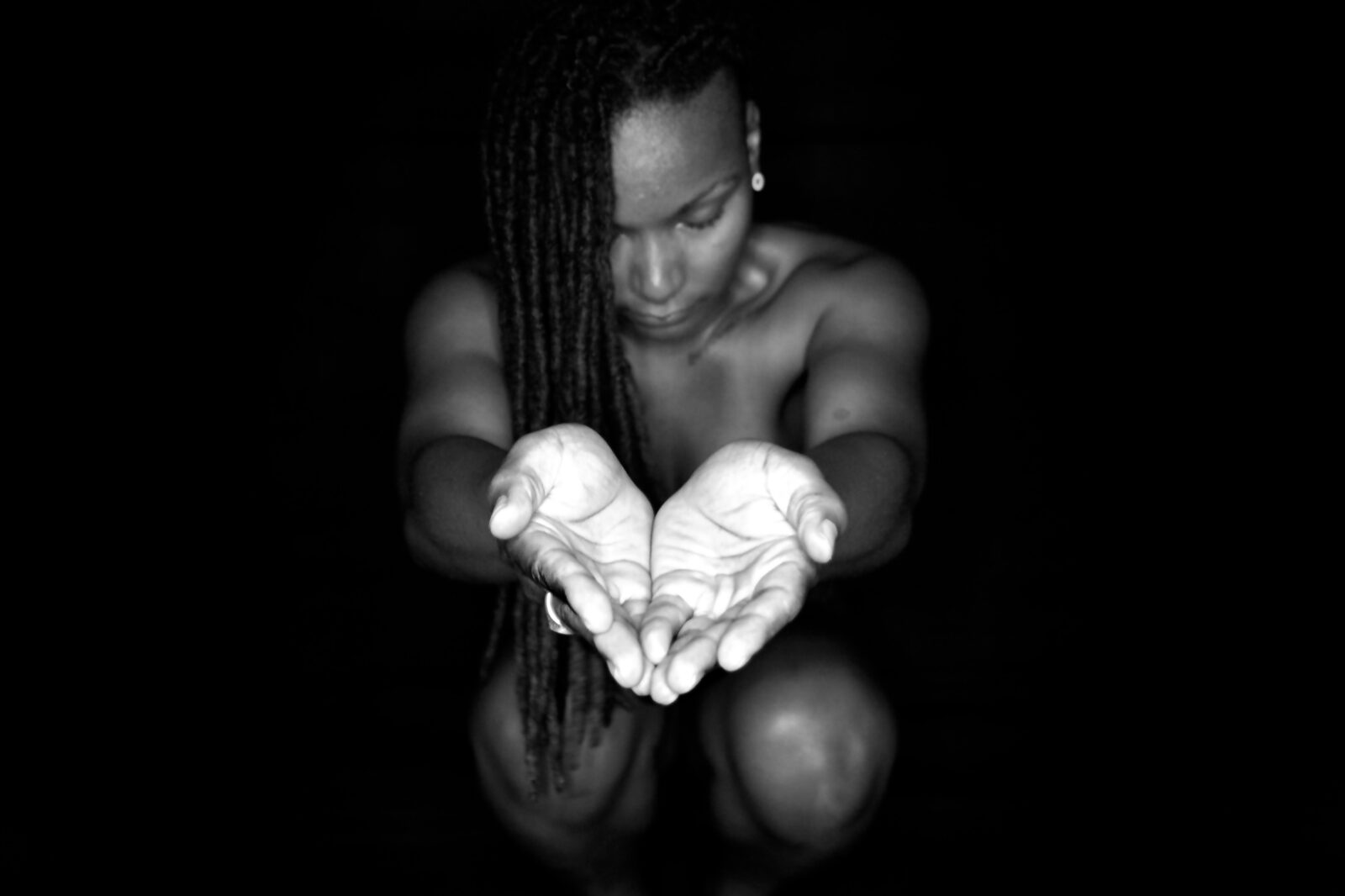 A black-and-white portrait showing a figure kneeling in darkness with long braided hair, gazing down at their own cupped hands extended toward the viewer and illuminated by a single light source against an otherwise black background.