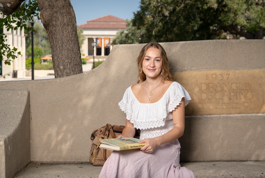 an occidental student sits on a bench with a book smiling on the los angeles campus.