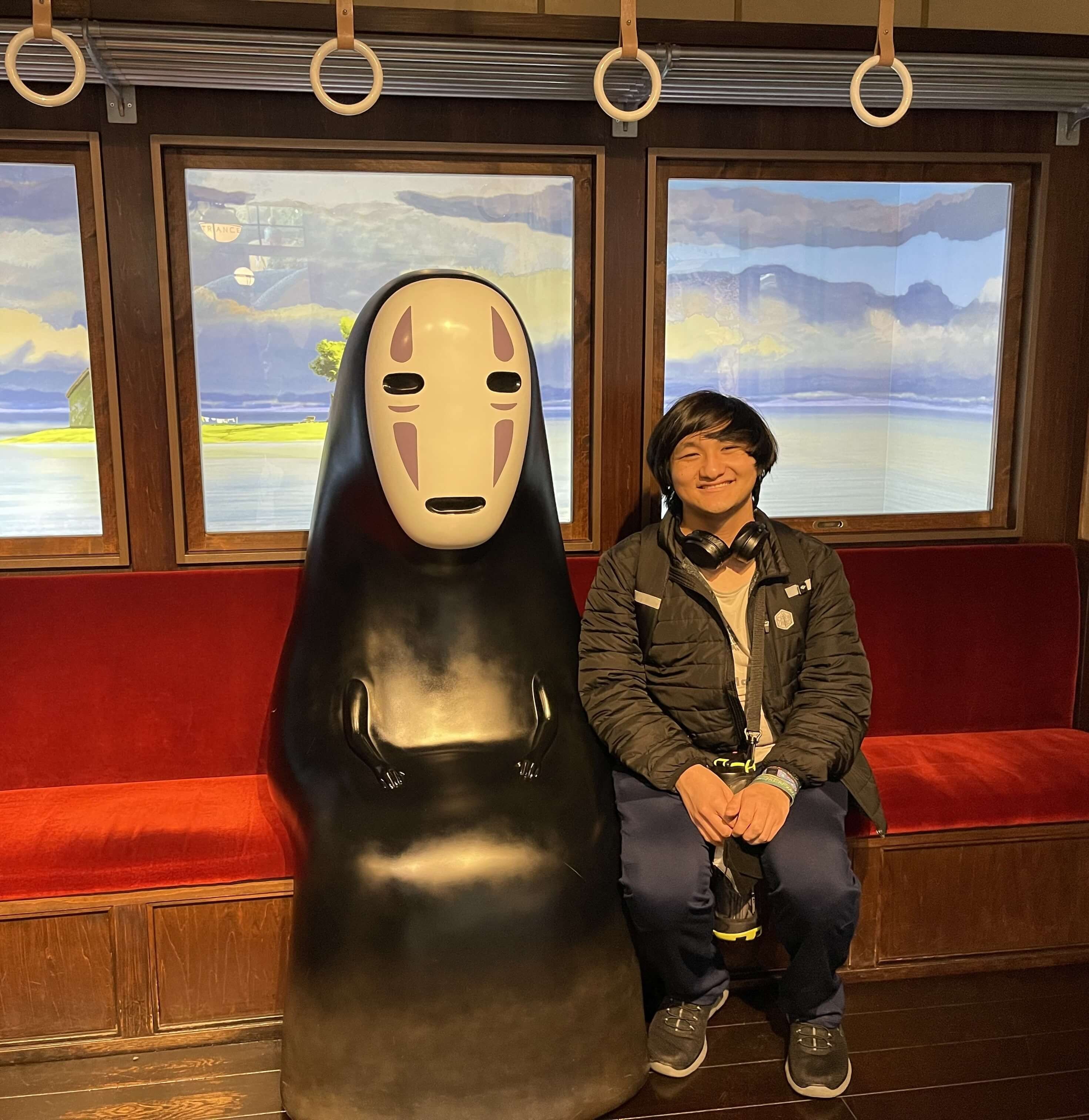 Photo of Zane sitting in a staged train interior next to Kaonashi (No-Face) from Hayao Miyazaki's film, Spirited Away.