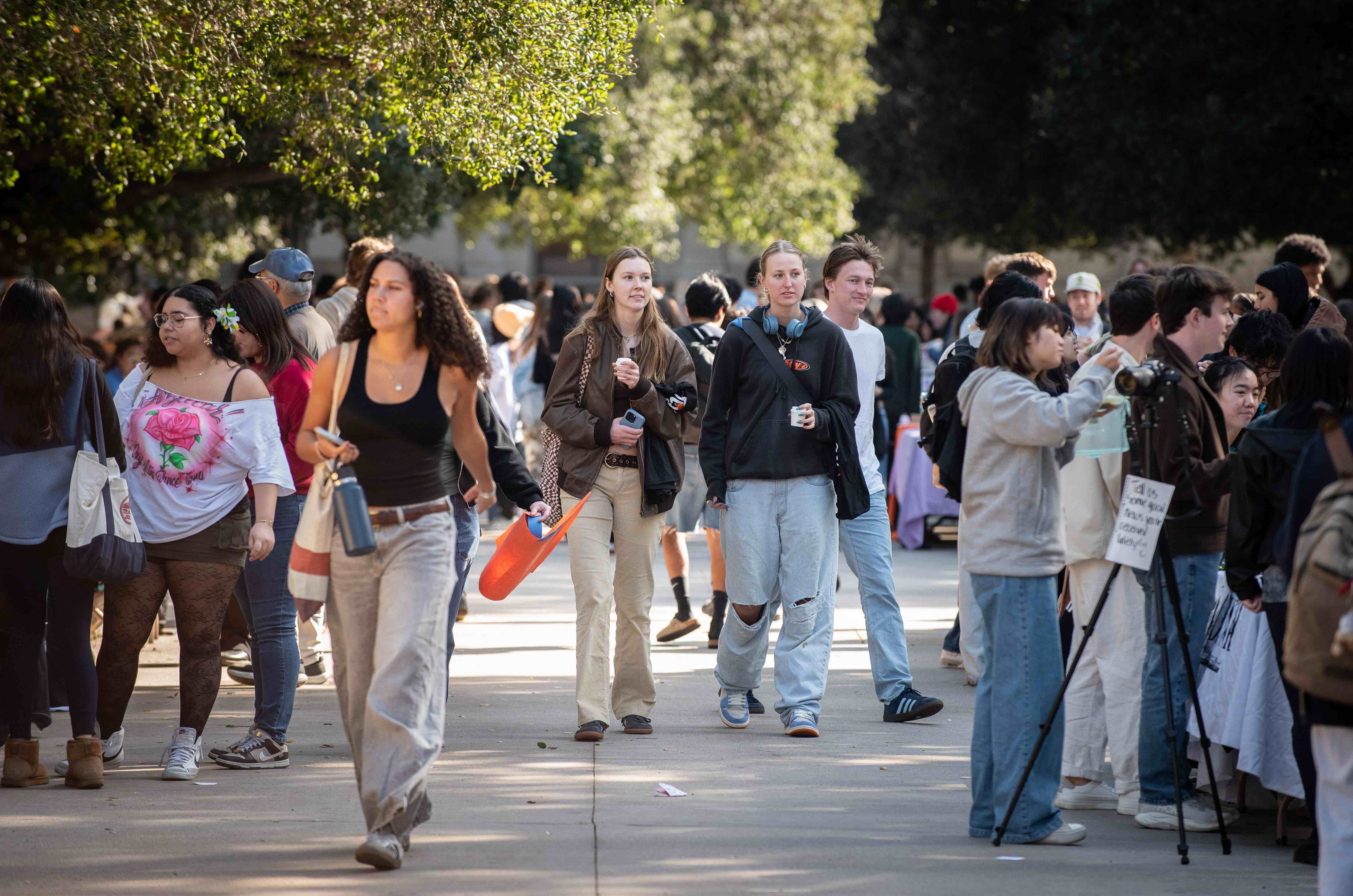 A bustling outdoor campus walkway filled with students, some walking and others stopping at tables for conversations. Trees with green foliage provide shade along the path. Students are casually dressed, carrying backpacks, water bottles, and tote bags.