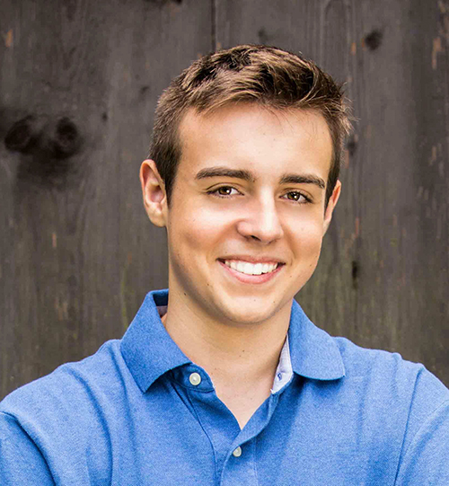 Braedon is wearing a blue polo shirt and is standing in front of a wood wall