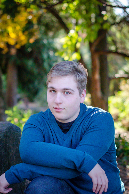 Zach is wearing a blue top and seated in front of a grove of trees