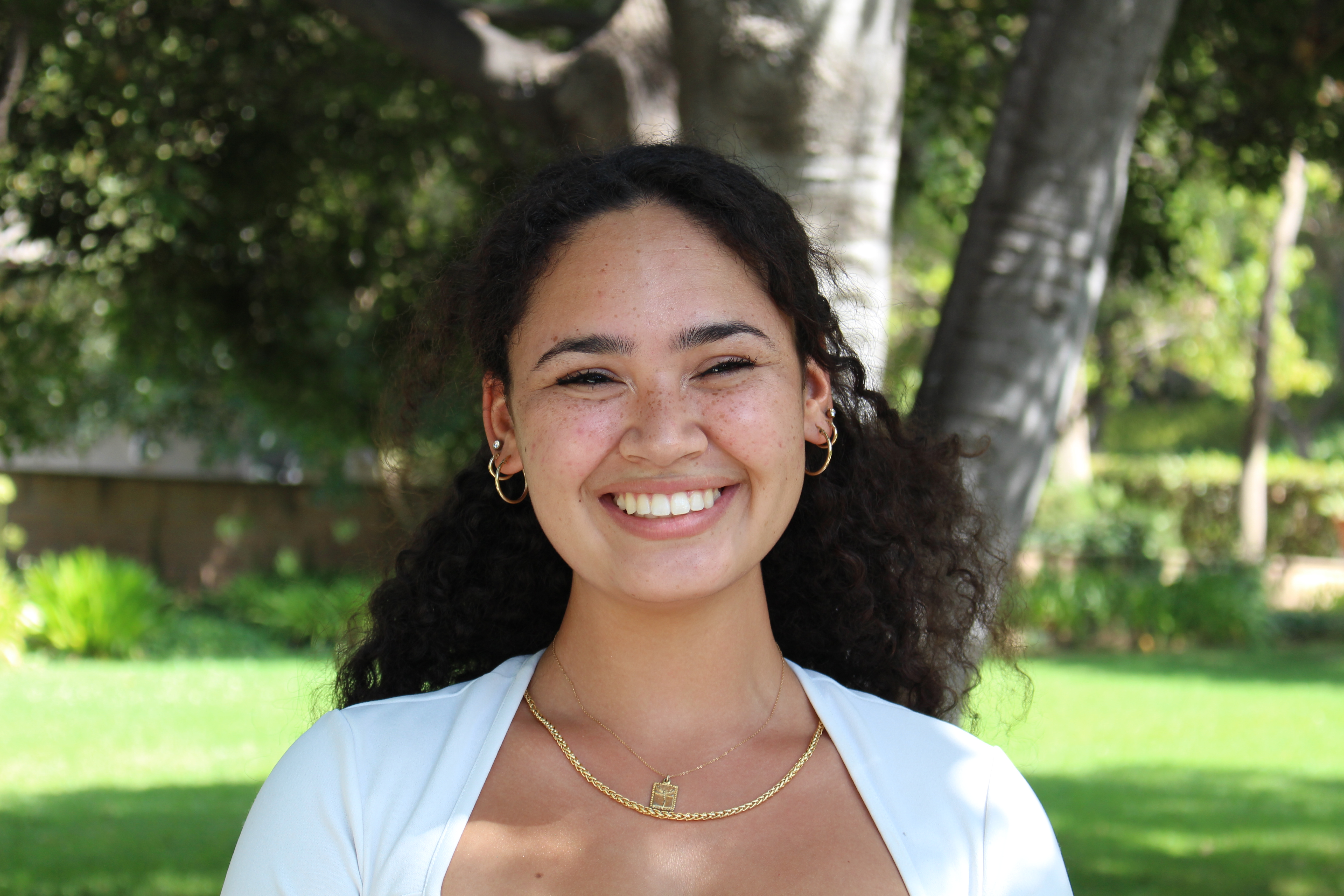 Carina smiles, wearing hoop earrings and a white cardigan, with trees and lawn in the background