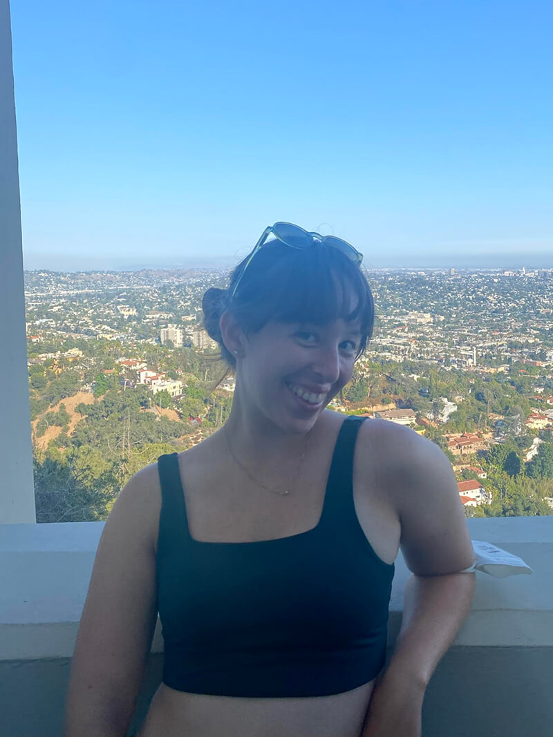 Emma is wering a black shirt and is standing in front of a window with a view of buildings and hills below