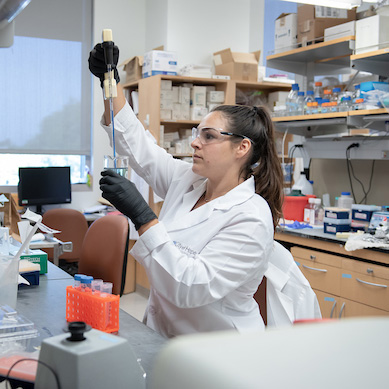 Young woman in lab coat and safety goggles uses pipette filled with blue liquid