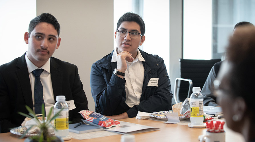 Two students in suits sitting at an office table listen attentively to a speaker