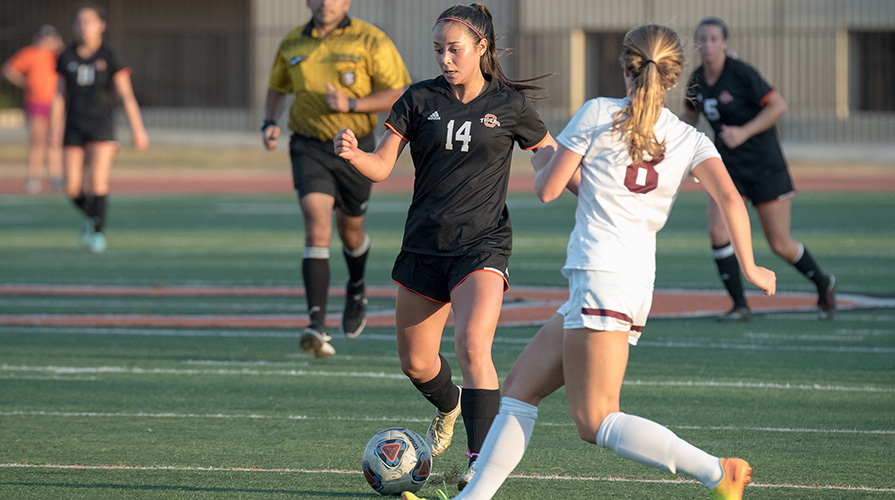Student wearing black uniform kicks soccer ball while her opponent runs to meet her