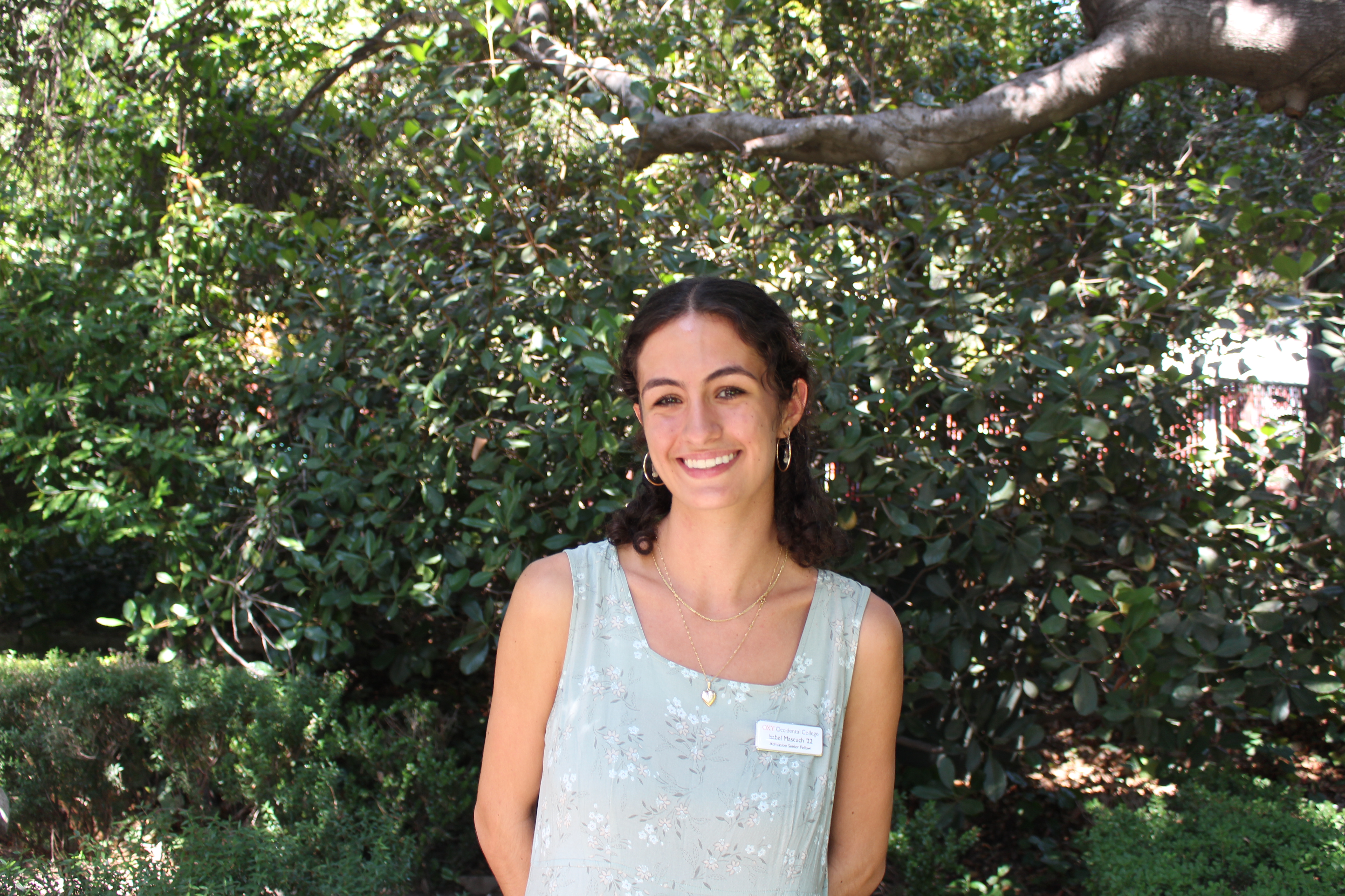 Isabel smiles, wearing a blue tank top and name tag, with trees in the background