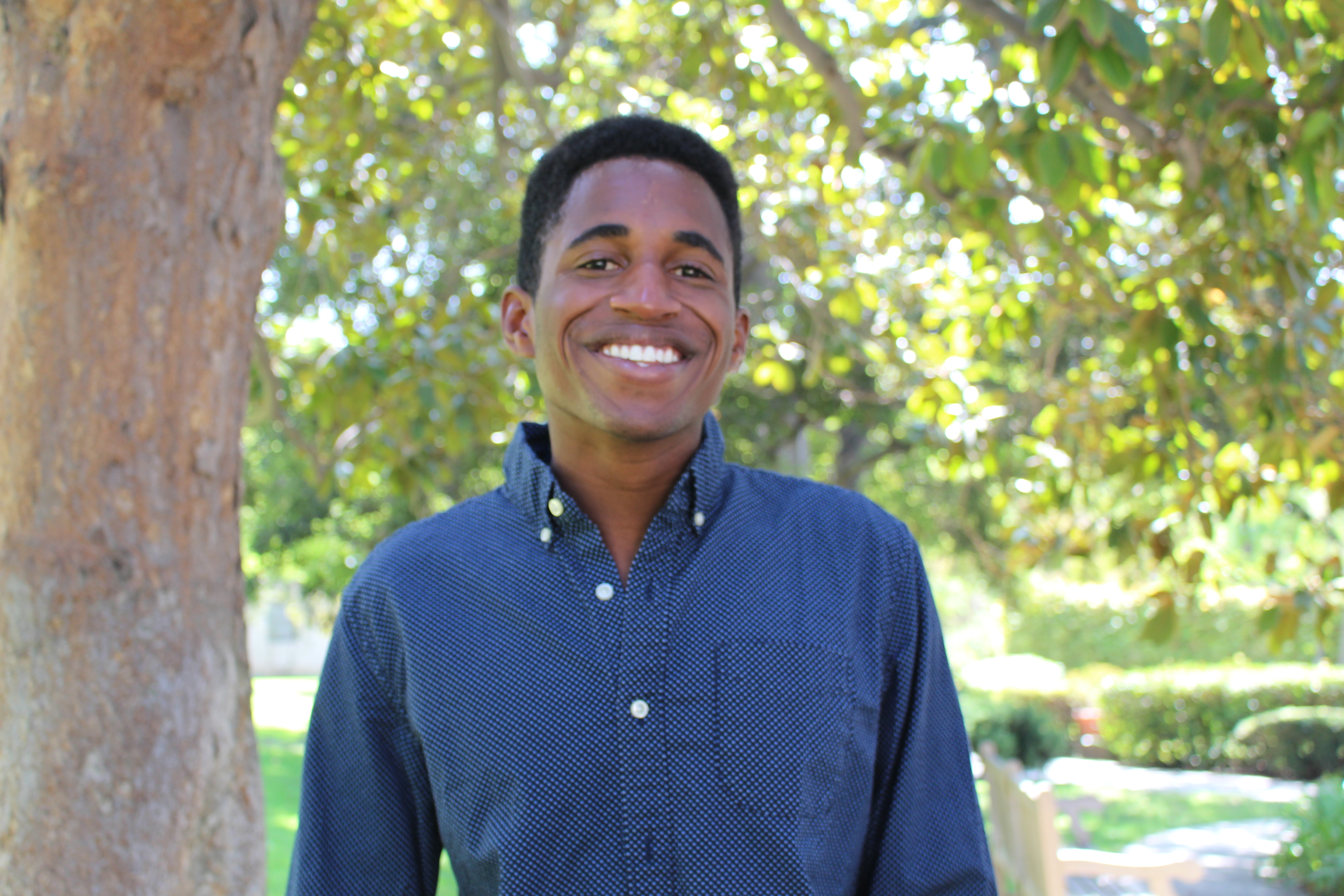 James smiles, wearing blue button up shirt with trees in the background