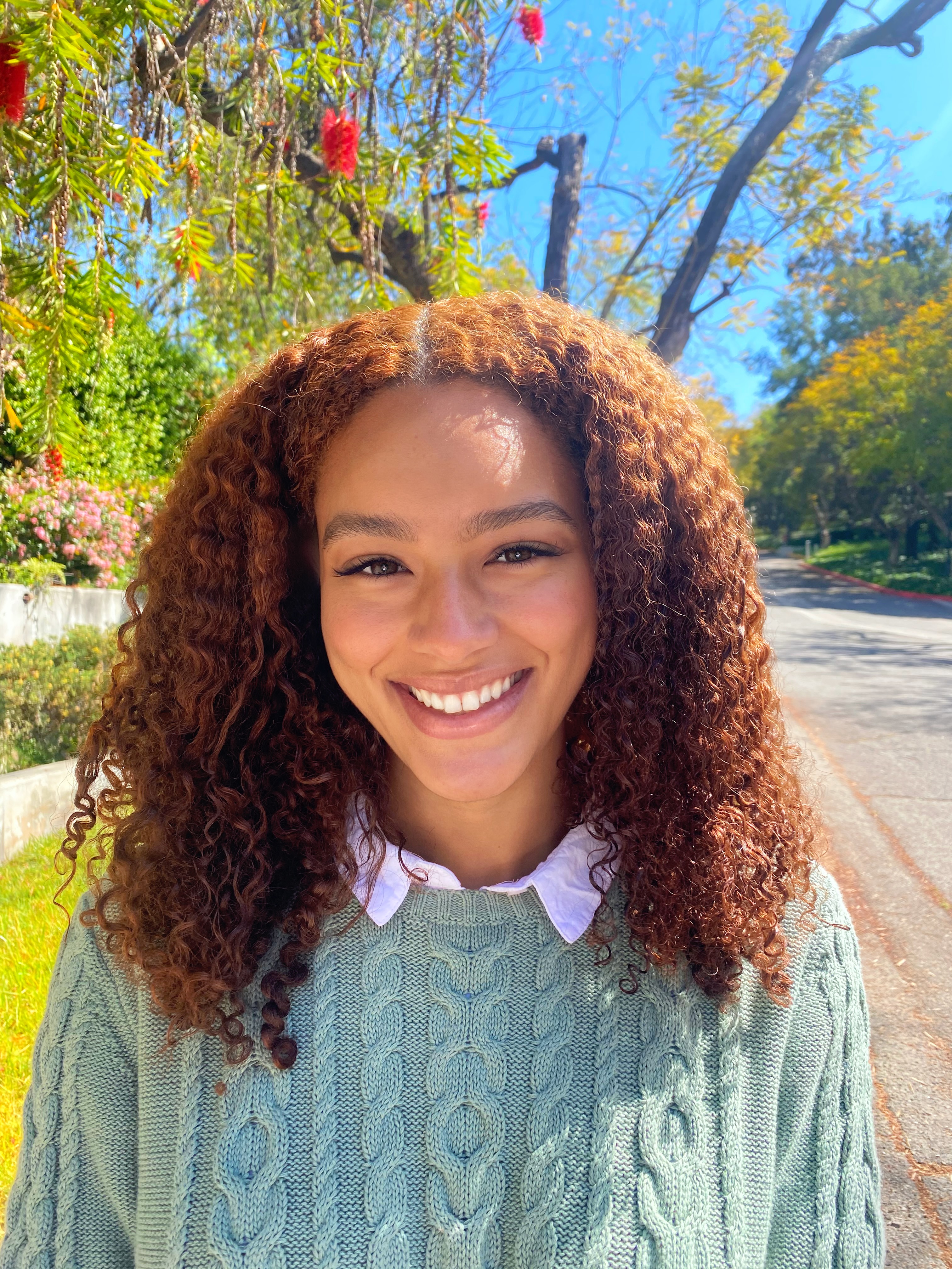 Kamea smiles, wearing a sage green sweater in front of a background of trees and blue sky