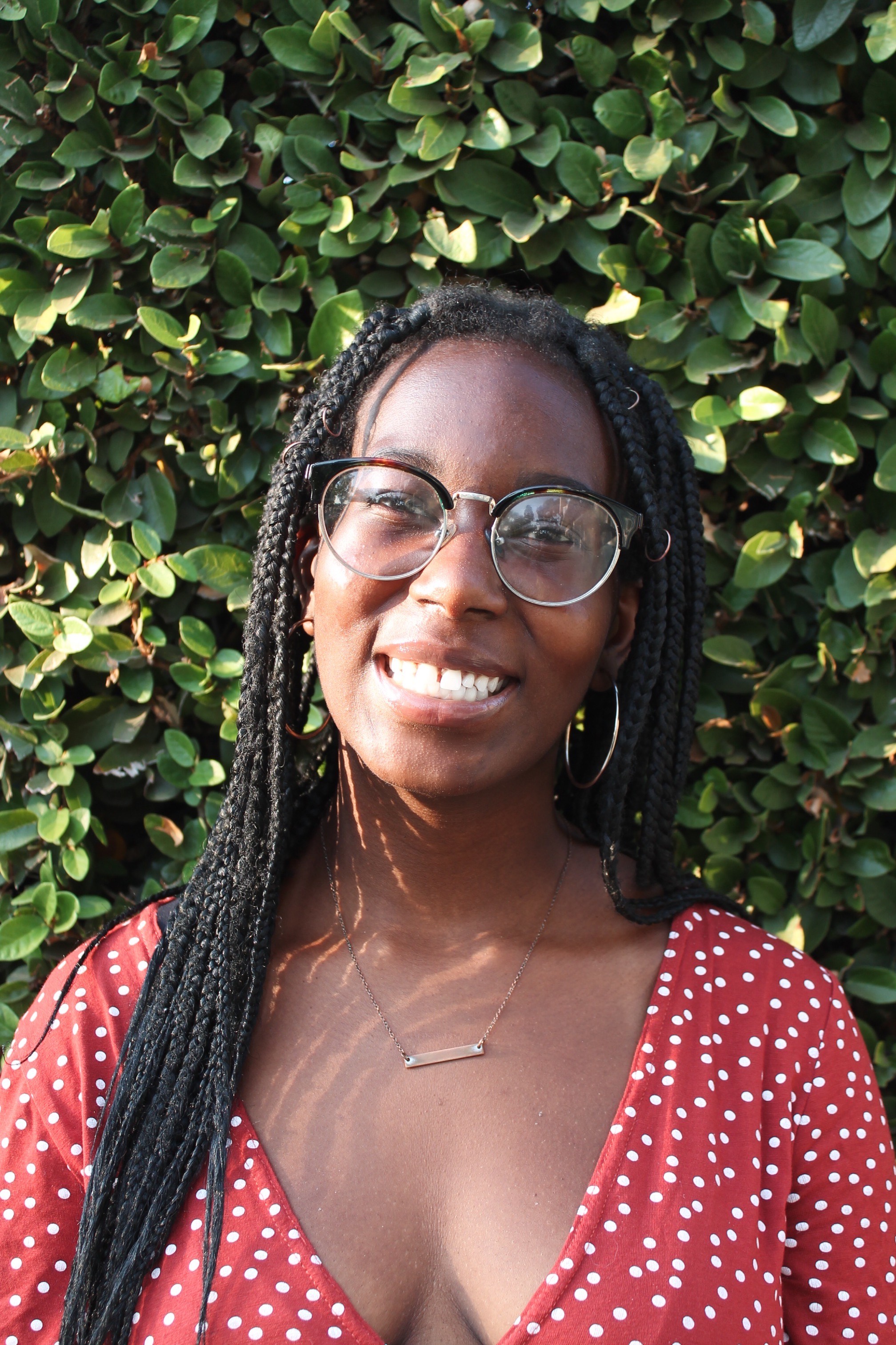 Katrina smiles, wearing glasses and a red and white polka dot dress, with foliage in the background