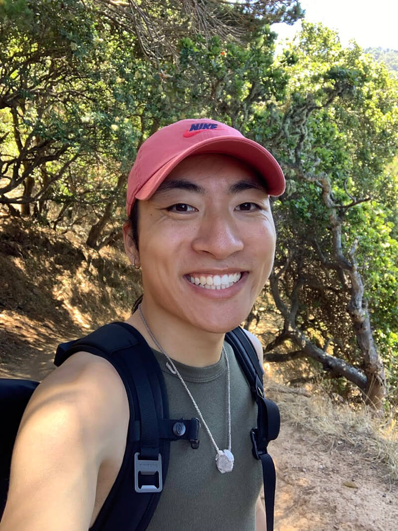Nathan is wearing a black shirt, backpack and a red hat with trees in the background