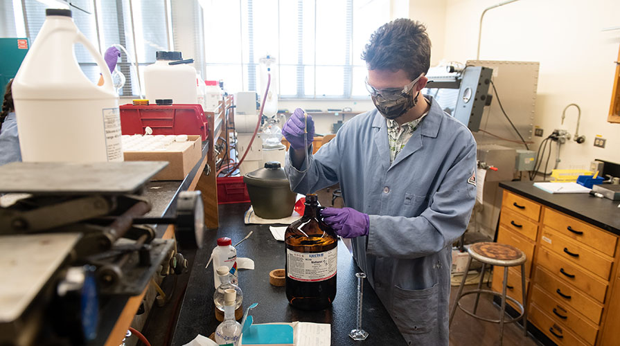 A student wearing a lab coat, gloves, and mask holds a beaker in a science lab