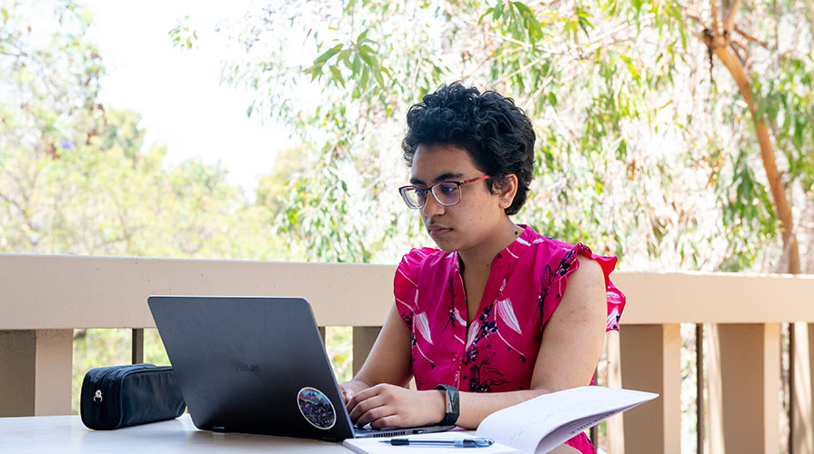  A student looks intently at a laptop in the foreground, with lush foliage in the background