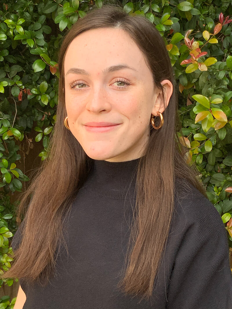 Sela is wearing a black shirt with greenery in the background