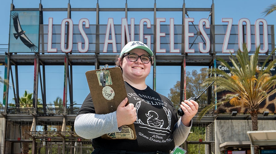 Oxy student intern with a clipboard in front of the Los Angeles Zoo sign