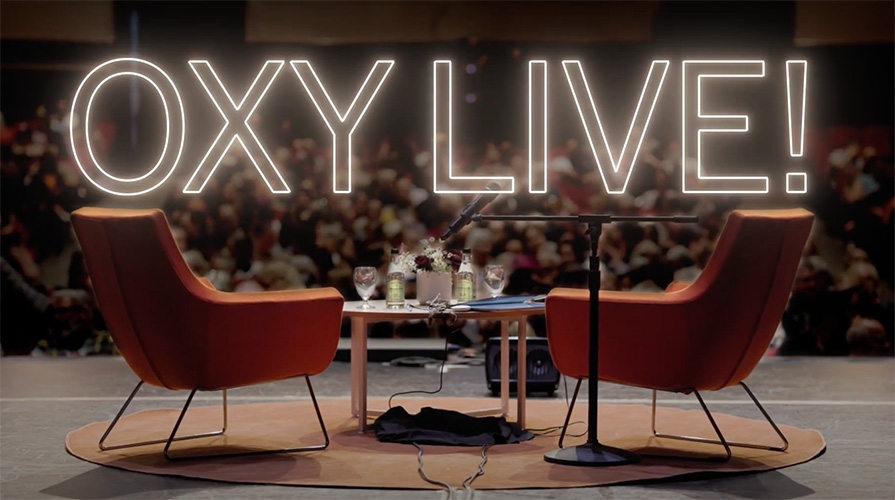 Two orange chairs on an empty stage with the header Oxy Live above