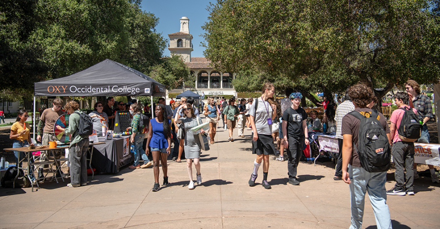  A bustling outdoor Involvement Fair at Occidental College. Students walk through a sunny campus plaza lined with club recruitment tables and an 'OXY' branded tent, showcasing a vibrant and active student life.
