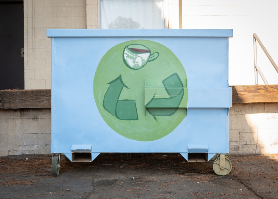 Dumpster painted with a blue background and green recycling "chasing arrows" symbol on it.