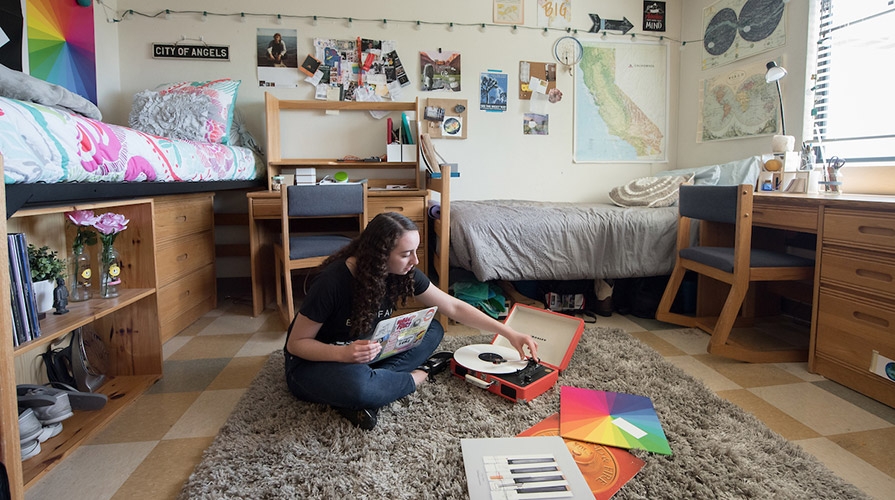 A student sits on the floor of her dorm room with a record player and a colorful selection of records