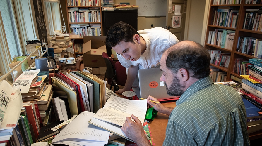 Oxy student Kevin Conroy leans attentively over a desk held by Professor Alexander Day in an office filled with books