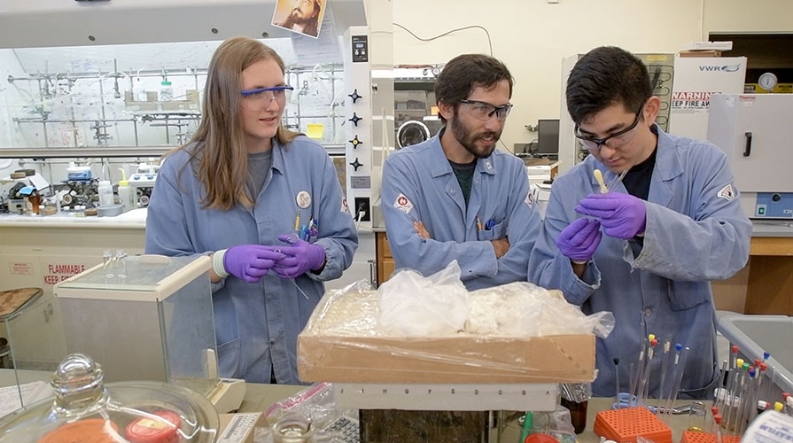 A professor and two students wearing lab coats, safety googles, and gloves hold beakers in a science lab