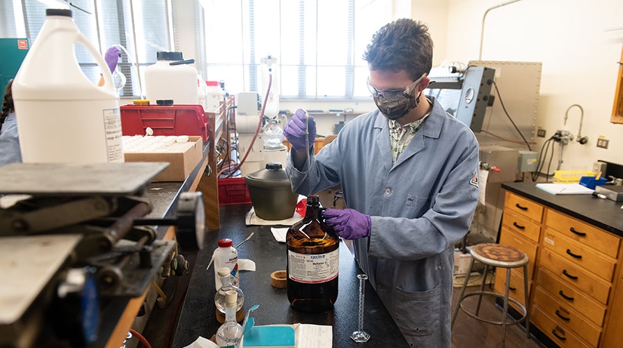 A student wearing a lab coat, gloves, and mask holds a beaker in a science lab