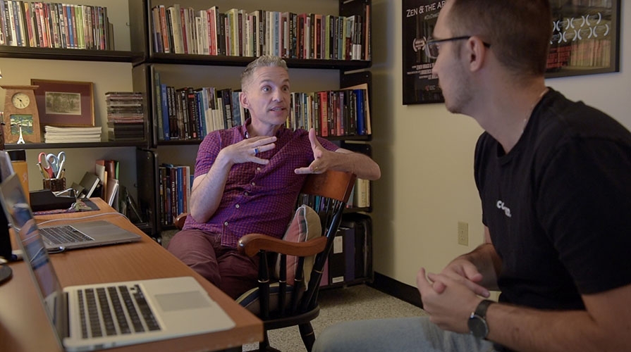 Faculty member Broderick Fox speaks to student Brendan Galbreath in an office full of books