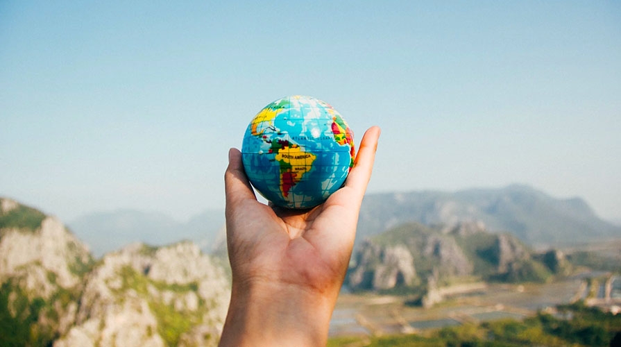 The foreground shows a hand holding a small globe, with mountains in the background