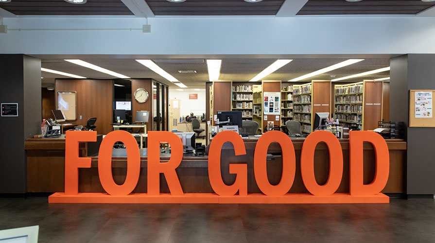 Large wooden orange sign reading "FOR GOOD" rests in front of a library circulation desk. 