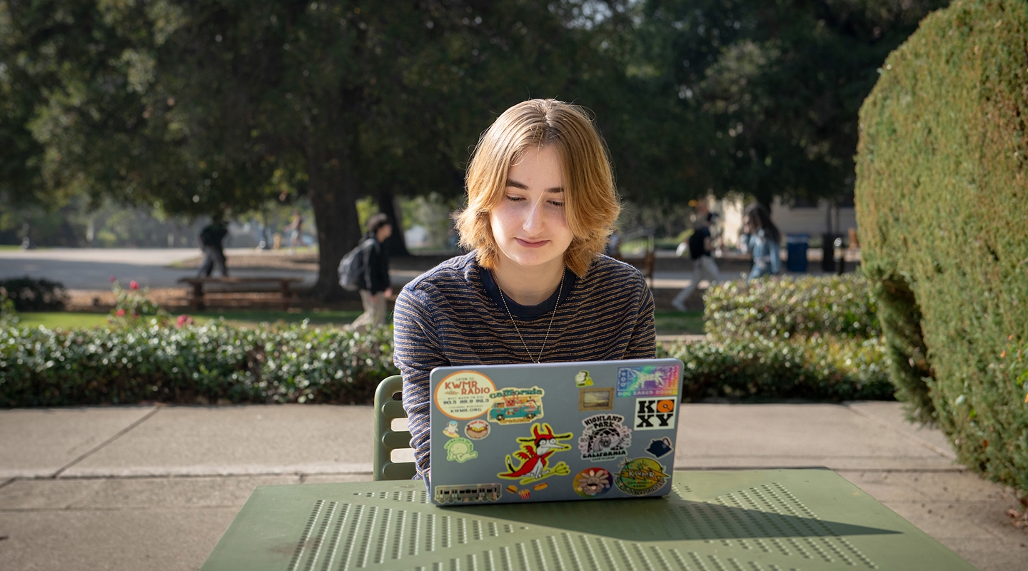 An Occidental College student sits at an outdoor table looking at her laptop.