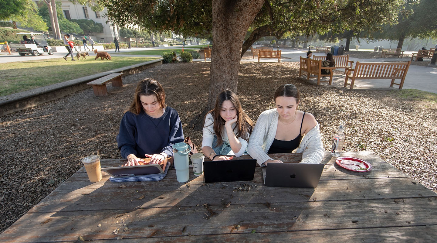 Three Occidental College students sitting side by side at a picnic table, looking at their laptops