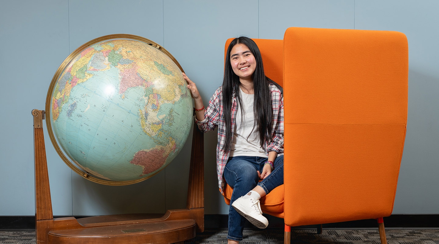 Thina Ly '27 sits next to a giant globe in the campus library, seated on an orange chair