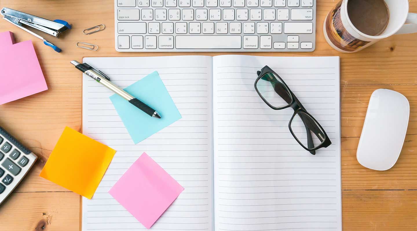 top-down view of an organized wooden office desk with various items including a keyboard, mug, notebook and colorful sticky notes